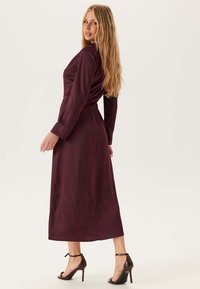 Abito midi bordeaux con maniche lunghe, tessuto morbido, vita aderente e uno spacco laterale, abbinato a sandali neri con tacco alto.