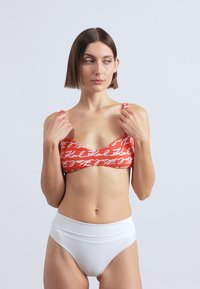 Czerwony top bikini z białym napisanym kursywą napisem, zestawiony z białymi, wysokim stanem. Gładka faktura, usztywniona konstrukcja, regulowane ramiączka.