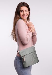 Graue Umhängetasche mit strukturierten Oberflächen, pinkem Reißverschlussakzent und verstellbarem Trageriemen. Modell trägt ein pinkes, geripptes Oberteil und helle Jeans.