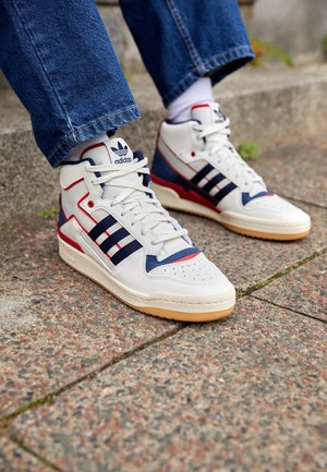 Białe, wysokie sneakersy Adidas z granatowymi pasami i czerwonymi akcentami, noszone z niebieskimi jeansami na zewnętrznych kamiennych schodach.