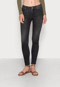 Fekete skinny farmernadrág sima textúrával, klasszikus öt zsebes dizájnnal és ezüst gombdíszítéssel. Barna lapos szandálokkal párosítva.