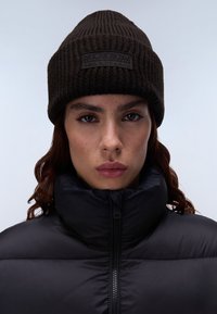 Berretto lavorato a maglia di colore marrone scuro con texture a costine, dotato di un patch logo nero. Indossato con un giubbotto imbottito nero con collo alto.