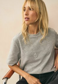 Chemise grise à manches courtes avec une coupe décontractée, en tissu doux. Présente un collier en argent avec des anneaux entrelacés. Pantalons sombres, posture assise.