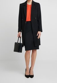 Černý dvouřadý kabát přes červenou halenku a černou sukni. Černé lodičky na vysokém podpatku a malá černá kabelka. Hladké textury a elegantní design.