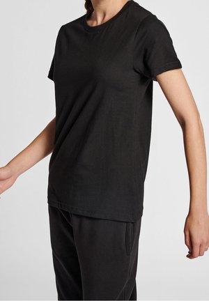 Person iført en ensfarvet sort t-shirt med korte ærmer og sorte bukser, stående mod en lys grå baggrund.