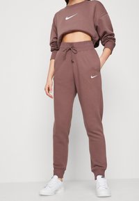 Person wearing mauve Nike sweatpants and cropped sweatshirt with white Nike sneakers, standing against plain white background.