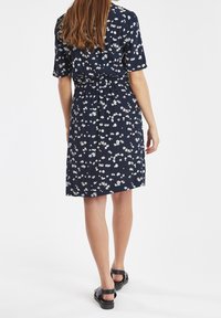 Kvinna stående med ryggen mot kameran, iklädd en marinblå blommig klänning och svarta sandaler mot en enkel vit bakgrund.