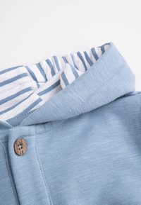 Jasnoniebieska bawełniana bluza z kapturem o teksturowanym wykończeniu, z białą i niebieską paskowaną podszewką oraz drewnianym guzikiem.