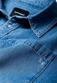 Koszula dżinsowa w jasnoniebieskim kolorze z ostrym kołnierzykiem. Posiada srebrny guzik oraz wyszywane inicjały "E.R." na szwie kieszeni.