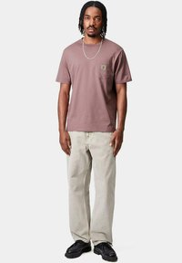 T-shirt in cotone marrone con taschino sul petto, abbinato a jeans grigi chiari e scarpe nere. Design semplice e rilassato con scollo rotondo.