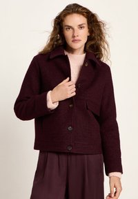 Woman with curly hair wearing a burgundy buttoned jacket over a light pink sweater and matching burgundy pants, standing against a plain background.