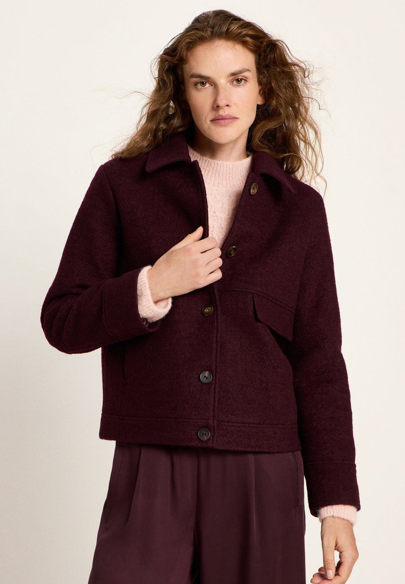 Woman with curly hair wearing a burgundy buttoned jacket over a light pink sweater and matching burgundy pants, standing against a plain background.