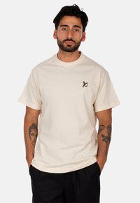 T-shirt in cotone crema a maniche corte, collo rotondo e una piccola grafica nella zona sinistra del petto. Vestibilità rilassata.