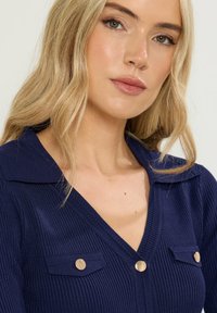 Blond kvinna med lätt smink som bär en marinblå ribbad tröja med guldknappar och bred krage, mot en ljus bakgrund.