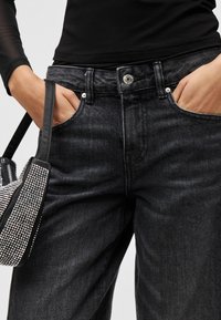 Osoba nosi čierne rifle s vypraným efektom, má ruky v vreckách a nesie čiernu kabelku ozdobenú striebornými trblietavými kamienkami.