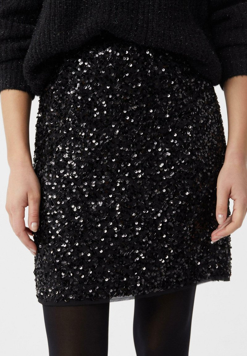 Schwarzer Paillettenrock mit figurbetontem Schnitt, glänzender Textur und schimmernder Oberfläche. Verfügbar oberhalb des Knies mit geradem Saum.