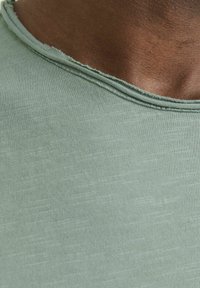 Light green cotton T-shirt with a textured surface, featuring a raw edge neckline and short sleeves, showing a close-up view of the fabric.