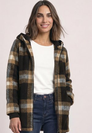 Femme portant une veste zippée à carreaux marron, noir et gris sur une chemise blanche et un jean bleu, souriante avec une main dans la poche.