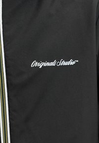 Crna jakna s bijelim tekstom "Originals Studio™" blizu prsa i patentnim zatvaračem sa zlatnim i bijelim rubovima sa strane.