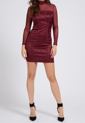 Donna che indossa un mini abito aderente color bordeaux con maniche lunghe trasparenti e tacchi alti neri con cinturino alla caviglia, in piedi davanti a uno sfondo semplice.