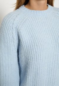 Gaiši zils adīts džemperis ar ribotu tekstūru, apaļu kaklu un raglan piedurknēm. Satur smalku kabelīša adījuma detaļu gar plecu šuvi.