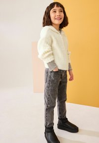 Maglione bianco morbido con zip e polsini a coste, foderato a righe all'interno, abbinato a jeans grigi e stivaletti neri. Posizione in piedi.