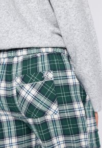 Groene, witte en marineblauwe geruite broek met een elastische tailleband en achterzak, gedragen met een lichtgrijze textuurtrui.