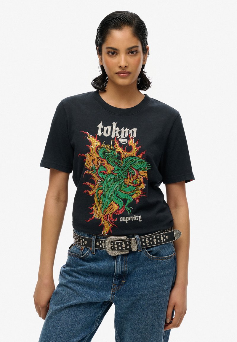 Zwarte katoenen T-shirt met een kleurrijke draak graphic, inclusief de tekst "Tokyo" en het "Superdry" logo, gecombineerd met blauwe spijkerbroek en een zwarte riem met studs.