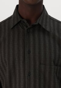 Ciemnozielona koszula zapinana na guziki z teksturowanym, paskowanym wzorem, klasycznym kołnierzykiem i kieszonką na piersi. Czarne guziki podkreślają design.