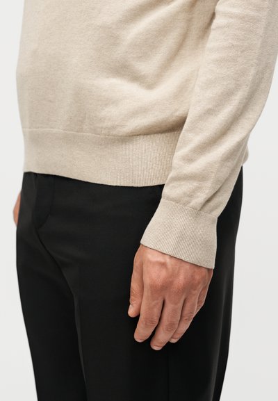 Μανσέτα μπεζ πουλόβερ με μακριά μανίκια που ακουμπά κοντά σε μαύρο, ραμμένο παντελόνι στη δεξιά πλευρά ενός ατόμου, δείχνοντας μέρος του χεριού και του καρπού τους.
