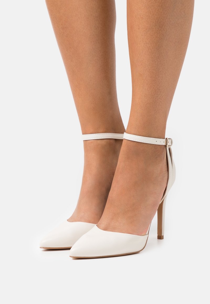 Jambes portant des escarpins blancs à bout pointu avec fines brides autour de la cheville et talons aiguilles sur un fond uni.