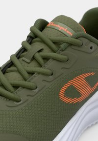 Olívazöld sneaker texturált anyaggal, kontrasztos narancssárga logóval és fehér talppal. Lapos cipőfűzőkkel és mintázott textúrával rendelkezik a felső részen.