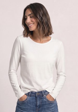 Femme aux cheveux bruns mi-longs portant une chemise blanche à manches longues et un jean bleu, souriante et regardant sur le côté, les mains dans les poches.
