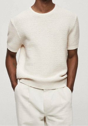 Homme portant un pull à manches courtes texturé de couleur crème, rentré dans un pantalon crème assorti, les mains dans les poches.