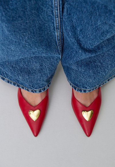 Pies con zapatos rojos de punta decorados con corazones dorados, combinados con jeans de mezclilla azul frente a un fondo claro.