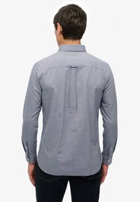 Mężczyzna w jasnoniebieskiej koszuli z długim rękawem, pokazany od tyłu, w ciemnych spodniach, na białym tle.