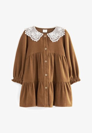 Brązowa sukienka z sztruksu z białym koronkowym kołnierzykiem, zapinana na guziki z przodu, z marszczonymi rękawami zakończonymi elastycznymi mankietami. Sukienka o wielowarstwowej konstrukcji z falbanką u dołu.
