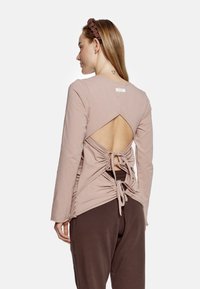 Długą rękawicę w miękkim beżowym materiale cechuje odsłonięte plecy z regulowanym wiązaniem oraz trójkątnym wycięciem. Połączona z brązowymi spodniami.