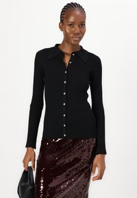 Schwarze gerippte Strickjacke mit Kragen und goldenen Knöpfen, kombiniert mit einem glitzernden burgunderroten Rock. Der Stoff wirkt weich und strukturiert.