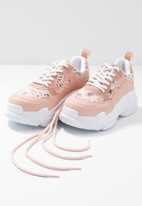 Baskets épaisses roses avec semelles blanches et lacets blancs, présentant un motif serpent beige et marron, exposées avec les lacets défaits sur une surface blanche.