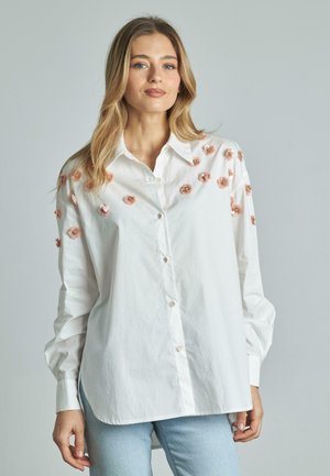Camicia bianca con bottoni, decorata con ricami floreali in rosa e oro, presenta maniche lunghe e un orlo curvo. Tessuto in cotone morbido.