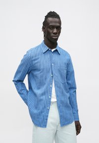 Mężczyzna z dredami w niebieskiej, pasiastym koszuli na guziki i jasnych dżinsach na jednolitym białym tle.