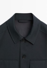 Camicia grigio scuro con colletto a punta, chiusura a un bottone nero e due tasche sul petto. Realizzata in tessuto liscio con un design strutturato.