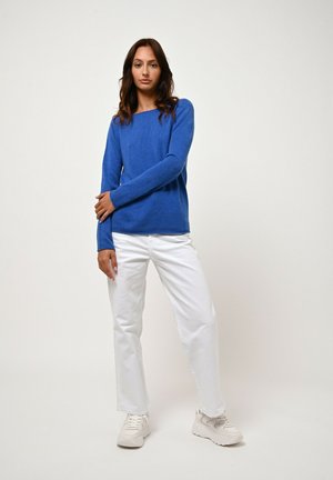 Vrouw die tegen een effen achtergrond staat in een blauwe sweater met lange mouwen, witte broek en witte sneakers, met één arm over haar lichaam gekruist.