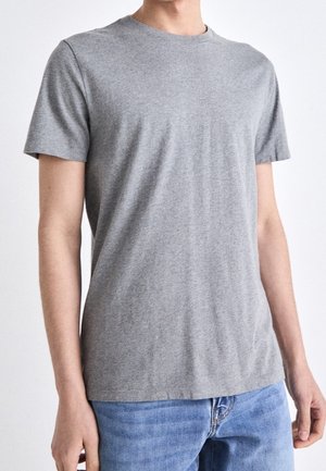 Młoda osoba ubrana w prosty, szary t-shirt z krótkim rękawem i niebieskie dżinsy, stojąca na tle jednolitego białego tła.