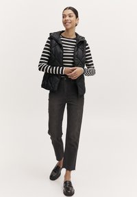 Chaleco acolchado negro con capucha sobre una camiseta de rayas en blanco y negro de manga larga, combinado con jeans oscuros de cintura alta y mocasines negros.