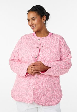 Naeratav tumedate juustega naine kannab roosat lillelist tikandiga jopet, millel on hõbedased trukid, seistes valge tausta ees.