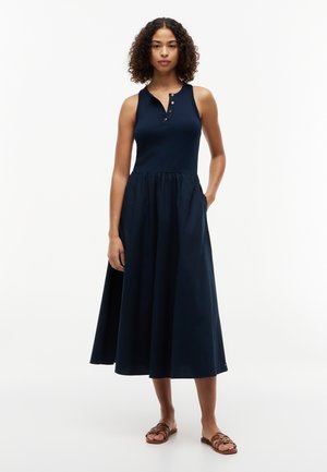 Kvinne med krøllete hår iført sleeveless navy midikjole med knappet halsutringning og brune flate sandaler, står med en hånd i lommen.