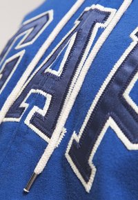 Tessuto blu con lettere cucite bianche e blu scuro e un cordino bianco con punta in metallo.