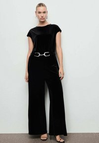 Mono de terciopelo negro con mangas cortas, piernas anchas y cintura ceñida con un cinturón que presenta una hebilla plateada. La modelo lleva zapatos de tacón negros con tiras.